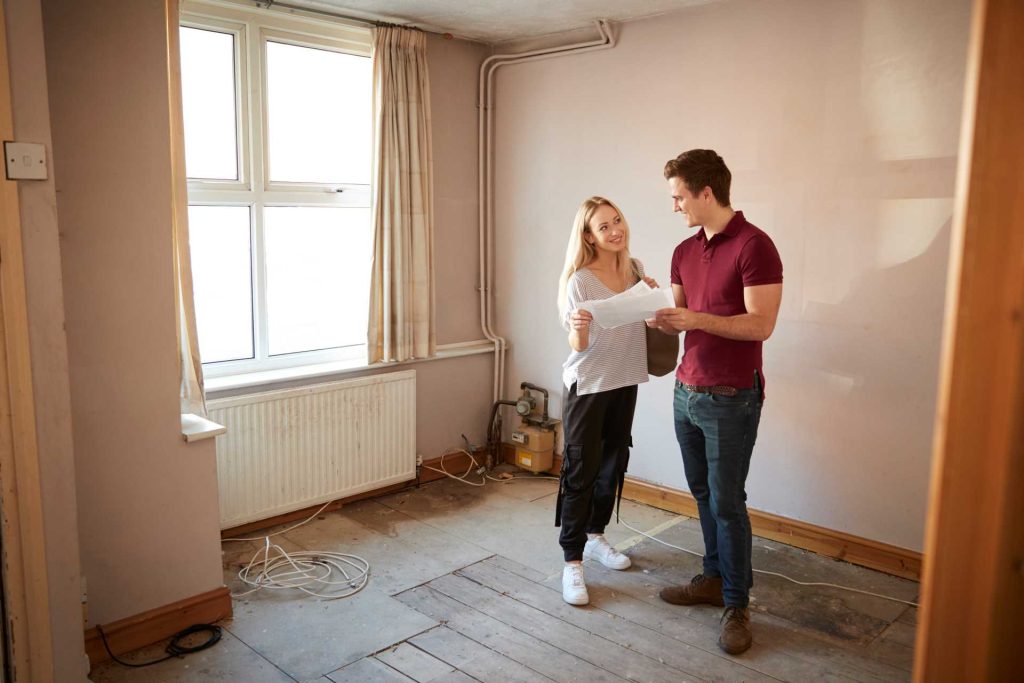 Young couple looking at building plans in a room undergoing renovations | Featured image for the blog How to Plan for a Home Renovation from BetaBoard.