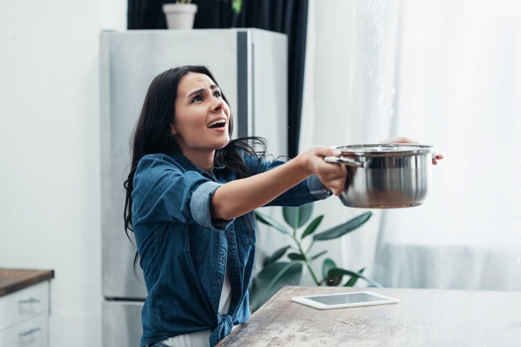 Worried lady catching water leaking from the ceiling in a cooking pot | Featured image for Water Damaged Ceiling Repair blog from BetaBoard.